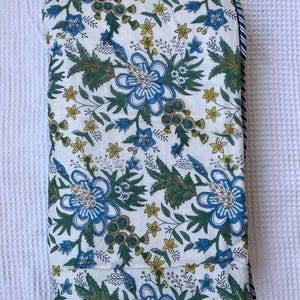 May include: Folded white fabric with a floral pattern featuring large blue flowers, green leaves, and small yellow blossoms. The edge has a blue and white striped border. The fabric is likely a quilt or blanket.