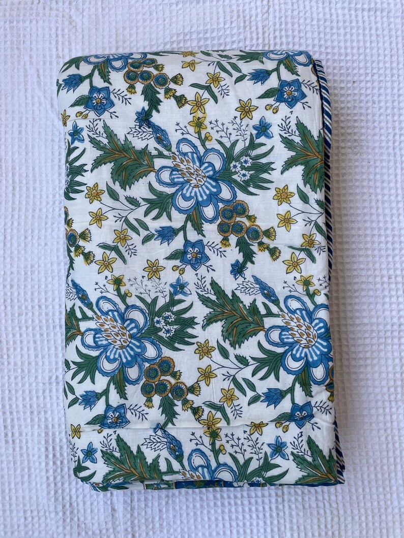 May include: Folded white fabric with a floral pattern featuring large blue flowers, green leaves, and small yellow blossoms. The edge has a blue and white striped border. The fabric is likely a quilt or blanket.