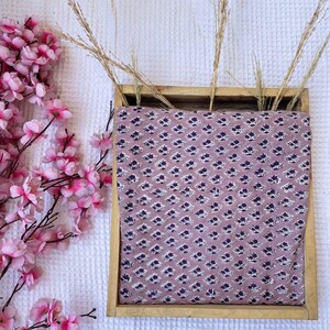 Puede incluir: Una bandeja de madera contiene tela con un estampado floral repetido en blanco y morado oscuro sobre un fondo malva. Hierbas secas y flores de cerezo rosas est&aacute;n dispuestas alrededor de la bandeja.