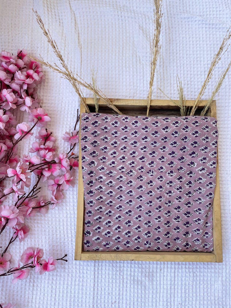 Puede incluir: Una bandeja de madera contiene tela con un estampado floral repetido en blanco y morado oscuro sobre un fondo malva. Hierbas secas y flores de cerezo rosas est&aacute;n dispuestas alrededor de la bandeja.