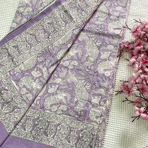 Op de afbeelding: Een lavendelkleurig textiel met een gedetailleerd wit vogel- en bloemenpatroon. De stof is gevouwen en toont een rand met een vergelijkbaar ontwerp. De randen zijn afgewerkt met een dunne, gestreepte bies. Roze bloemen staan op de achtergrond.