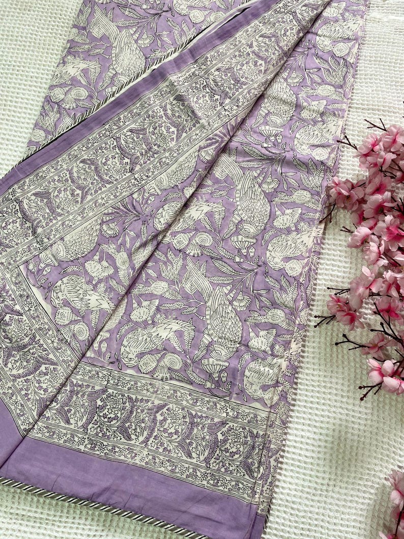 Op de afbeelding: Een lavendelkleurig textiel met een gedetailleerd wit vogel- en bloemenpatroon. De stof is gevouwen en toont een rand met een vergelijkbaar ontwerp. De randen zijn afgewerkt met een dunne, gestreepte bies. Roze bloemen staan op de achtergrond.