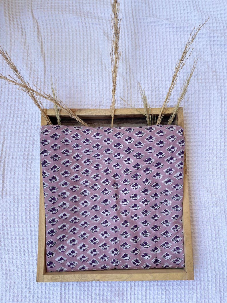 Puede incluir: Una bandeja de madera contiene una tela malva con un estampado floral azul oscuro repetido. Altas hierbas secas se alzan desde la bandeja. El fondo es una superficie blanca texturizada.