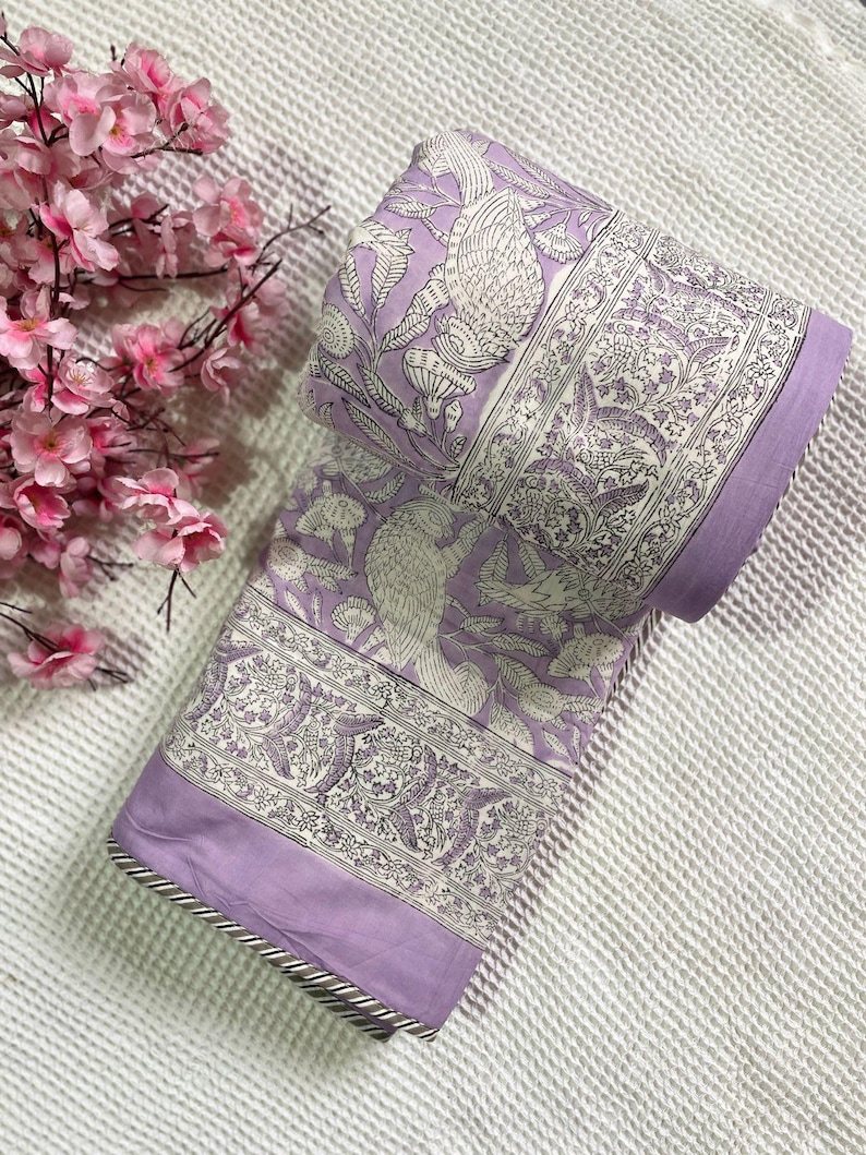 Op de afbeelding: Gerolde lavendelkleurige stof met een gedetailleerd wit bloemen- en vogelpatroon. De stof heeft een rand met een zilveren en grijze afwerking. Roze bloemen bevinden zich in de linkerbovenhoek.