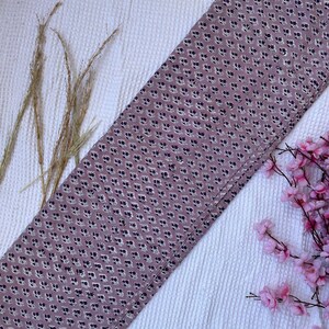 Puede incluir: Un trozo rectangular de tela con un patr&oacute;n floral repetido en azul oscuro y blanco sobre un fondo malva. La tela est&aacute; doblada y descansa sobre una superficie blanca texturizada, con hierba seca y flores rosas en el fondo.