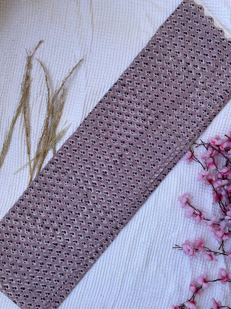 Puede incluir: Un trozo rectangular de tela con un patr&oacute;n floral repetido en azul oscuro y blanco sobre un fondo malva. La tela est&aacute; doblada y descansa sobre una superficie blanca texturizada, con hierba seca y flores rosas en el fondo.