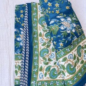 May include: A folded blue and white floral quilt with a repeating pattern of large flowers and leaves. The quilt has a border of white and green floral designs and a solid blue edge. The quilt is folded on a white surface.