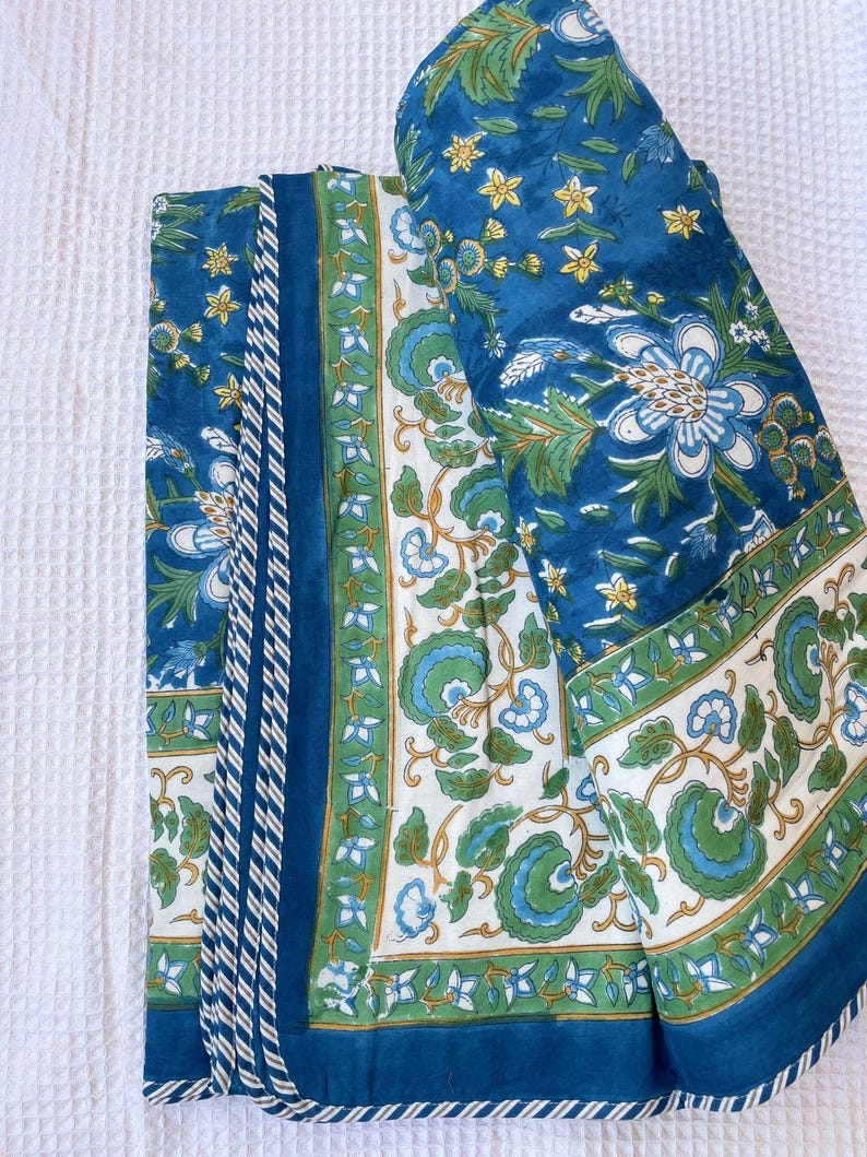 May include: A folded blue and white floral quilt with a repeating pattern of large flowers and leaves. The quilt has a border of white and green floral designs and a solid blue edge. The quilt is folded on a white surface.