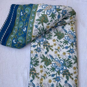 May include: A folded, floral-patterned quilt featuring a white background with blue and green flowers and foliage. The quilt's border is a deep blue with a green and white design. The quilt is folded, showcasing its intricate pattern and color scheme.