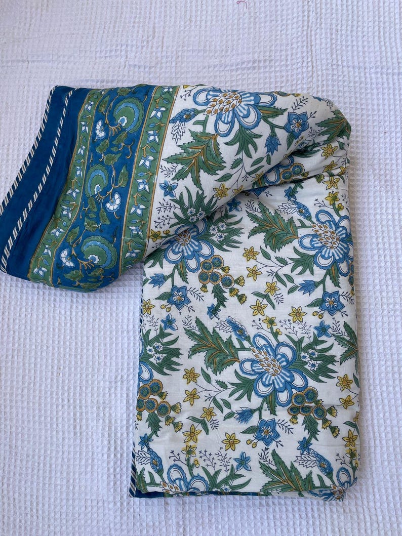 May include: A folded, floral-patterned quilt featuring a white background with blue and green flowers and foliage. The quilt's border is a deep blue with a green and white design. The quilt is folded, showcasing its intricate pattern and color scheme.