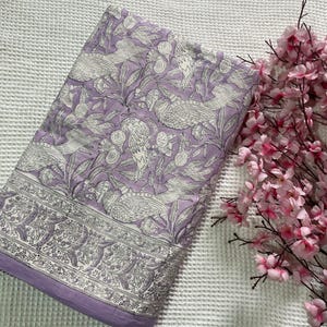 Op de afbeelding: Een gevouwen lavendelkleurige quilt met een wit vogel- en bloemenpatroon. De quilt heeft een decoratieve rand en een effen lavendelkleurige rand. Roze bloemen staan op de achtergrond.