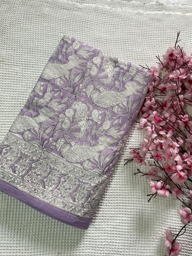 Op de afbeelding: Een gevouwen lavendelkleurige quilt met een wit vogel- en bloemenpatroon. De quilt heeft een decoratieve rand en een effen lavendelkleurige rand. Roze bloemen staan op de achtergrond.
