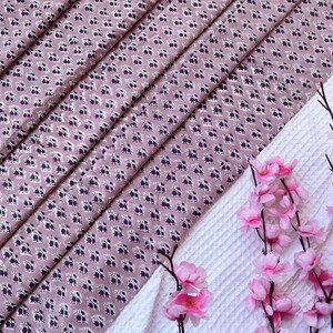 Puede incluir: Primer plano de tela plegada con un patr&oacute;n floral en tonos malva, blanco y morado oscuro. Un tejido blanco texturizado y flores rosas en el fondo. El patr&oacute;n floral se repite.