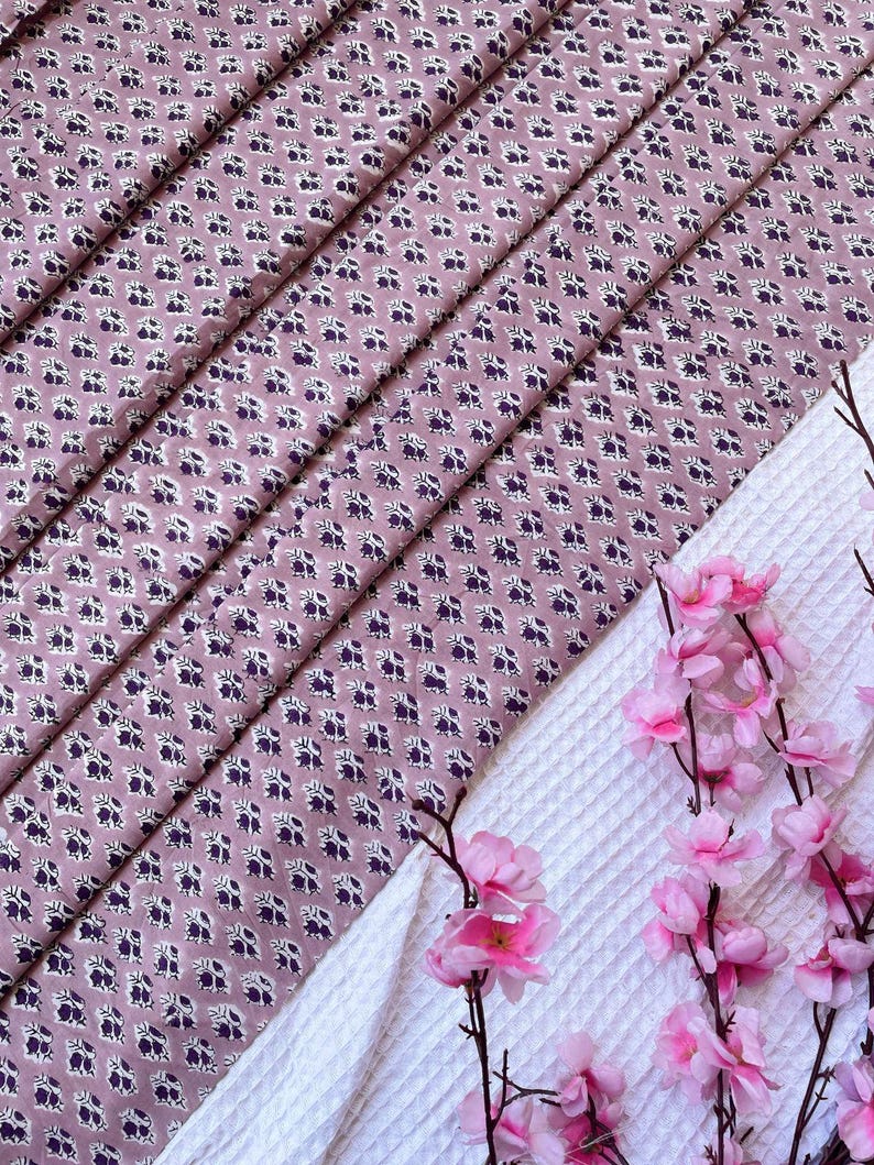 Puede incluir: Primer plano de tela plegada con un patr&oacute;n floral en tonos malva, blanco y morado oscuro. Un tejido blanco texturizado y flores rosas en el fondo. El patr&oacute;n floral se repite.