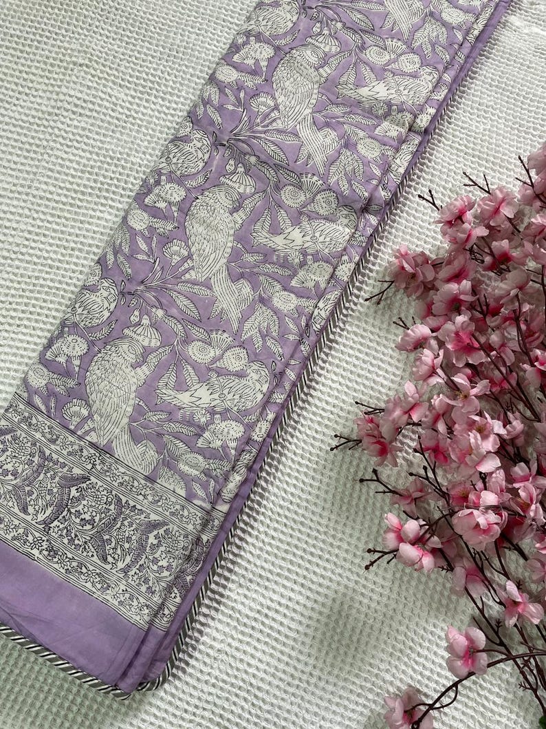 Op de afbeelding: Een lavendelkleurige gewatteerde stof met een wit uilen- en bloemenpatroon. De stof heeft een decoratieve rand en is opgevouwen. Roze bloemen staan op de achtergrond.