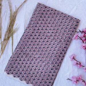 Puede incluir: Tela doblada con un estampado floral repetido en tonos malva y azul marino. La tela tiene un borde festoneado en color crema. Se muestra sobre una superficie blanca texturizada.