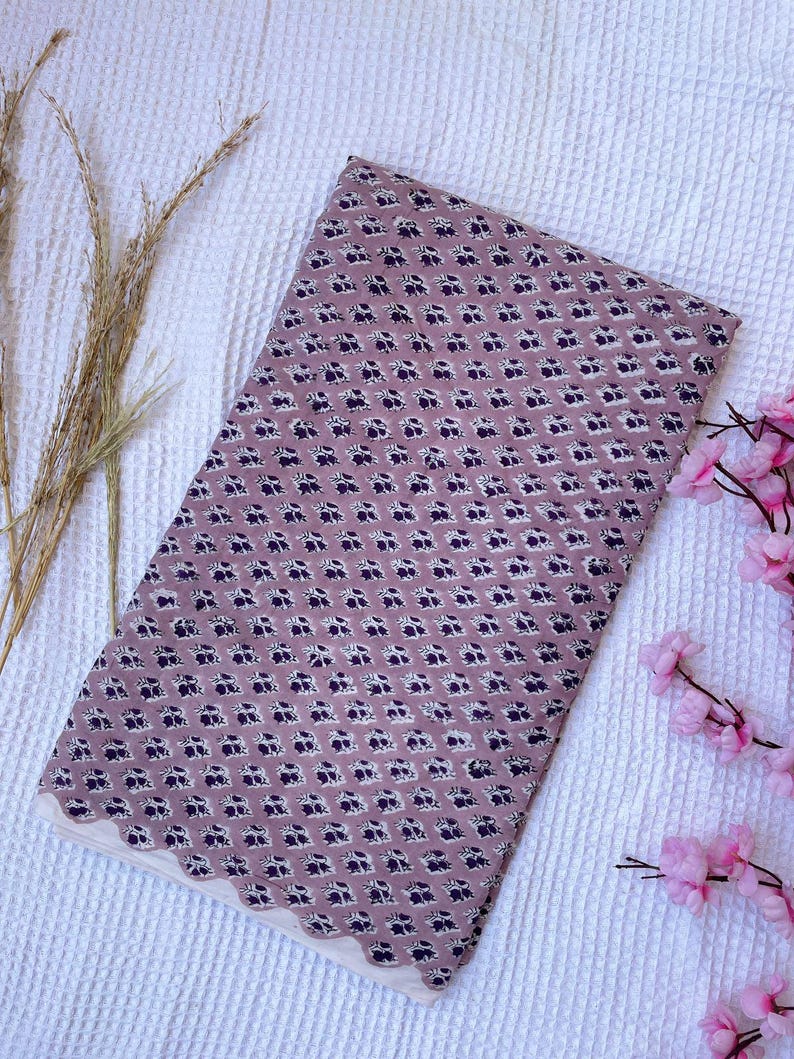 Puede incluir: Tela doblada con un estampado floral repetido en tonos malva y azul marino. La tela tiene un borde festoneado en color crema. Se muestra sobre una superficie blanca texturizada.