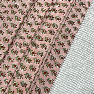 May include: Pink fabric with a repeating floral pattern in red and green. The fabric is folded, revealing the pattern and texture. A white, textured fabric is visible on the right side of the image.