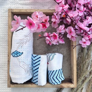 May include: Three white cotton baby swaddles with blue and white patterns, folded and displayed in a wooden box with pink flowers on top.