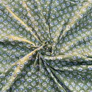 May include: Olive green fabric featuring a repeating pattern of white and light blue floral motifs. The fabric is gathered, creating folds and shadows. The texture appears soft and slightly wrinkled, with a detailed floral design.