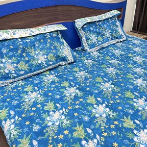 May include: Blue floral patterned bed set with two pillowcases. The bedding features a repeating pattern of white flowers, green leaves, and yellow star-shaped accents on a blue background. The pillowcases have a white and green border.