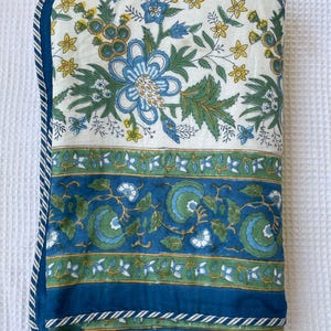 May include: A folded floral quilt featuring a white background with blue and green floral patterns. The quilt has a blue border with a white and blue striped trim. The design includes blue flowers, green leaves, and yellow accents.