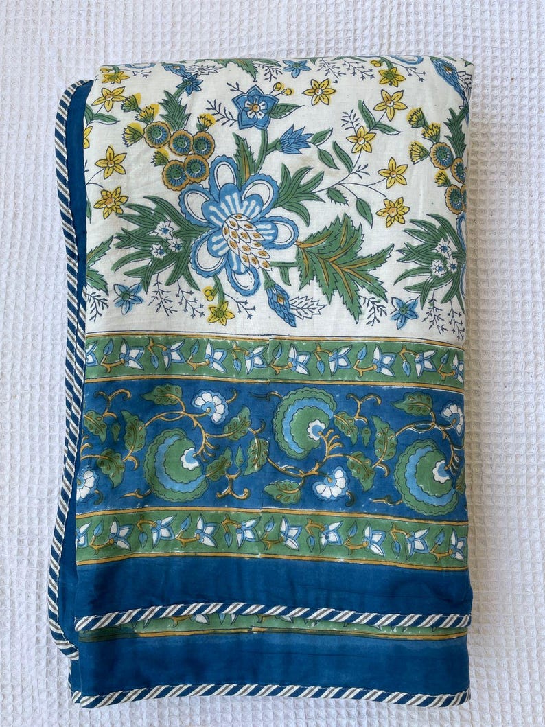 May include: A folded floral quilt featuring a white background with blue and green floral patterns. The quilt has a blue border with a white and blue striped trim. The design includes blue flowers, green leaves, and yellow accents.