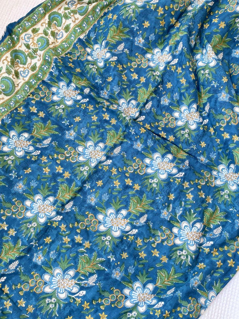 May include: A blue fabric with a floral pattern. The design features large white and light blue flowers, green leaves, and small yellow star-shaped accents. A border with a green and white floral pattern is visible.
