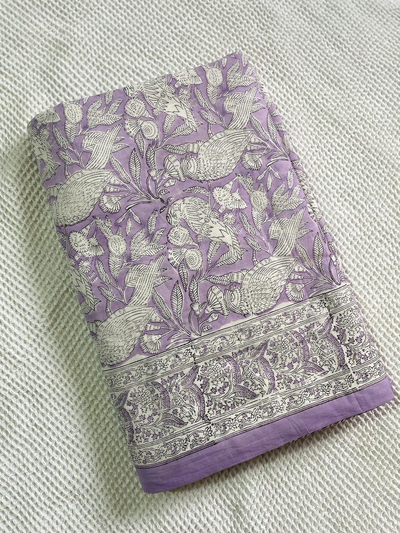 Op de afbeelding: Gevouwen lavendelkleurige stof met een wit vogel- en bloemenpatroon. De stof heeft een rand met een gedetailleerd ontwerp. De achtergrond is een wit, getextureerd oppervlak.