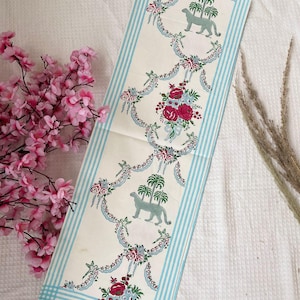 Puede incluir: Una tela rectangular blanquecina con un patrón repetido de rayas azul claro, guirnaldas florales y leopardos verdes estilizados. Los elementos florales incluyen rosas rojas y rosas. La tela es probablemente un camino de mesa decorativo.