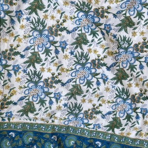 May include: White fabric with a repeating floral pattern in shades of blue, green, and yellow. The design features large stylized flowers and smaller blossoms, with a border of blue and green floral motifs. The fabric has a soft, slightly wrinkled appearance.