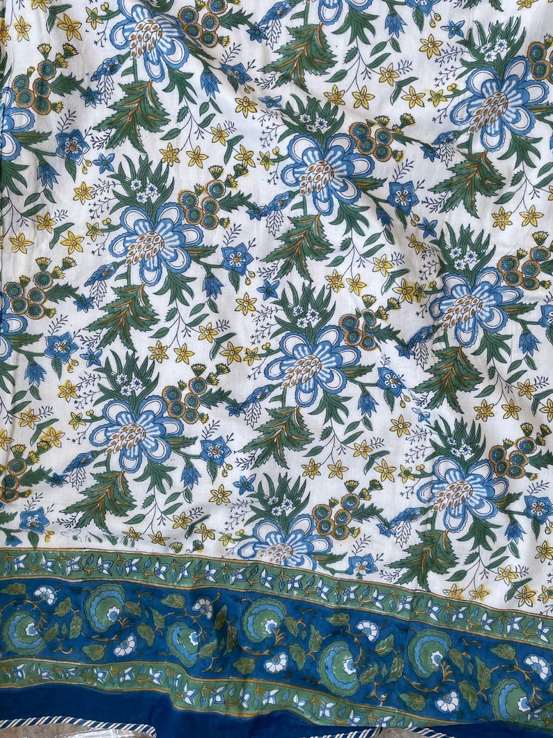 May include: White fabric with a repeating floral pattern in shades of blue, green, and yellow. The design features large stylized flowers and smaller blossoms, with a border of blue and green floral motifs. The fabric has a soft, slightly wrinkled appearance.