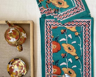 Op de afbeelding: Een set van turquoise placemats met een bloemenontwerp met oranje en gele bloemen, witte bladeren en een rood-witte geometrische rand. Een goud-rode bloemen theepot en schotel staan in een houten dienblad.