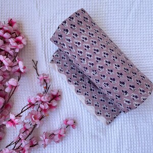 Puede incluir: Tela doblada con fondo lavanda y un estampado floral morado oscuro repetido. La tela est&aacute; colocada sobre una superficie blanca texturizada, con flores rosas en la esquina.