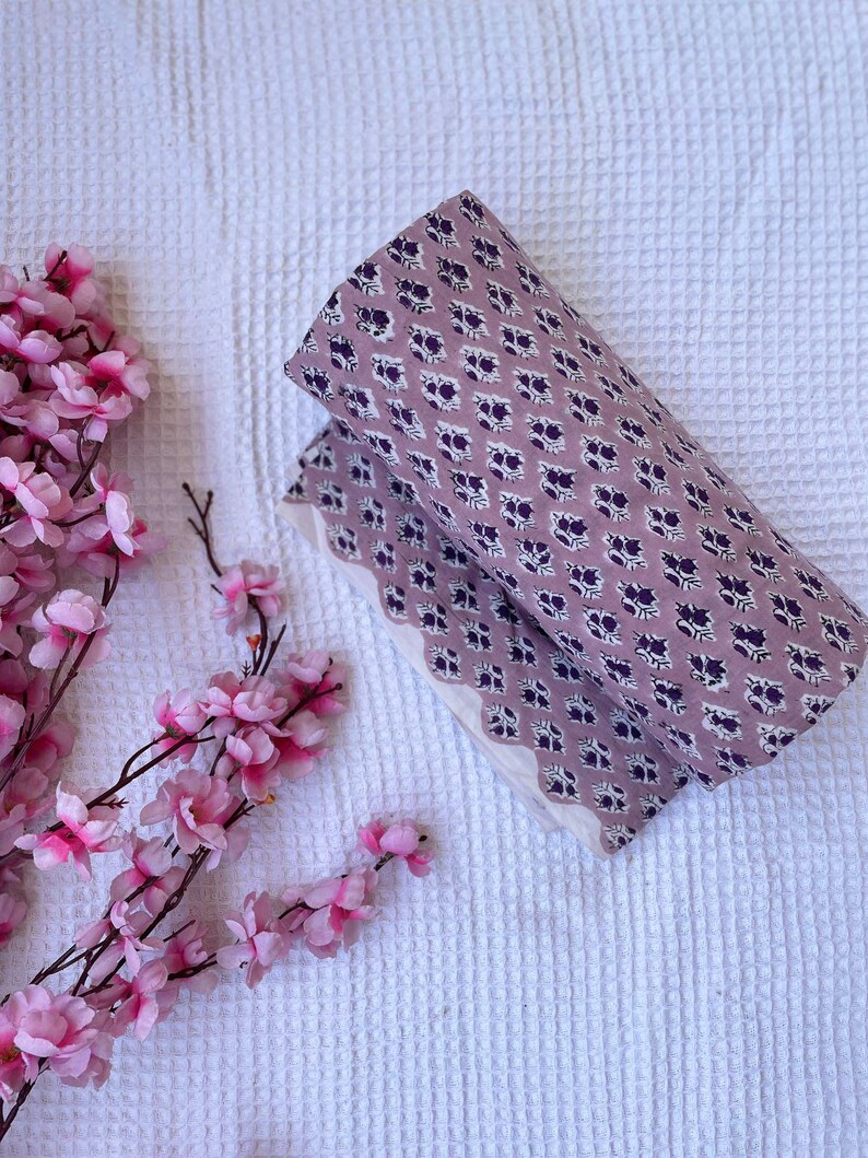 Puede incluir: Tela doblada con fondo lavanda y un estampado floral morado oscuro repetido. La tela est&aacute; colocada sobre una superficie blanca texturizada, con flores rosas en la esquina.