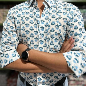 May include: White button-down shirt with a blue floral pattern. The shirt has long sleeves and a chest pocket. The floral design is repeated across the fabric. The shirt is worn with a black watch and a black belt.