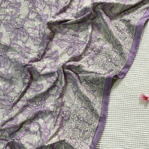 Op de afbeelding: Een gedrapeerde lavendelkleurige stof met een gedetailleerd wit bloemen- en vogelpatroon. De stof heeft een rand met een vergelijkbaar ontwerp. De plooien en schaduwen cre&euml;ren diepte en benadrukken de ingewikkelde print. Het thema is textielontwerp.