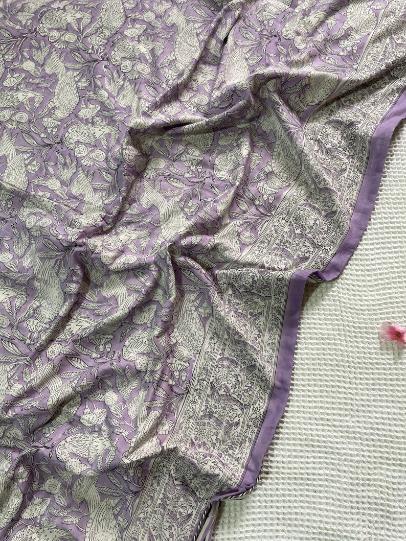 Op de afbeelding: Een gedrapeerde lavendelkleurige stof met een gedetailleerd wit bloemen- en vogelpatroon. De stof heeft een rand met een vergelijkbaar ontwerp. De plooien en schaduwen cre&euml;ren diepte en benadrukken de ingewikkelde print. Het thema is textielontwerp.