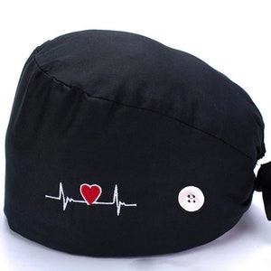 Puede incluir:  gorro de quirófano negro con un corazón rojo y un gráfico de latido del corazón bordado en la parte delantera. El gorro tiene un botón blanco en el lateral y una corbata en la parte posterior.