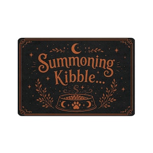 Peut inclure: Un paillasson rectangulaire noir avec le texte orange Summoning Kibble. Le motif comprend un croissant de lune, des étoiles, des branches feuillues et un graphique de gamelle pour animaux avec une empreinte de patte au centre en bas.