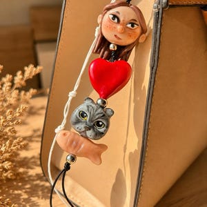 Puede incluir: Un colgante para bolso con una figura esculpida de una persona con cabello castaño y pecas, un corazón rojo brillante, una cara de gato gris y un pequeño pez color melocotón, unidos por cordones negros y blancos con cuentas metálicas plateadas.