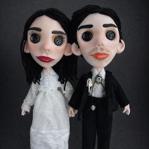 May include: Two dolls with black yarn hair and black button eyes. The left doll wears a white lace dress with long sleeves. The right doll wears a black suit jacket, black trousers, a white collared shirt, and a black bolo tie with a silver accent.