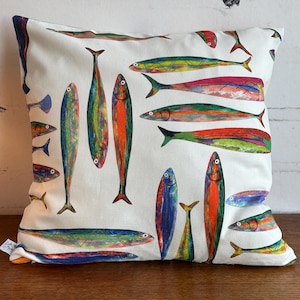 Könnte beinhalten: Quadratisches Kissen mit weißem Hintergrund und einem bunten Fischmuster. Die Fische sind in verschiedenen Größen und Ausrichtungen dargestellt, mit leuchtenden Farben wie Rot, Grün, Blau und Gelb. Das Kissen ist wahrscheinlich aus einem weichen Stoff.