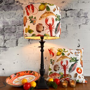 Könnte beinhalten: Eine schwarze Lampe mit Lampenschirm und Kissen mit Meeresfrüchtedesign. Der Lampenschirm und das Kissen haben einen weißen Hintergrund mit Illustrationen von Hummer, Fisch, Zitronen und Muscheln. Eine ovale Servierplatte und Kerzen sind ebenfalls vorhanden.