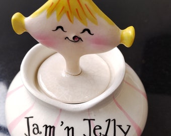 May include: White ceramic jar with pink stripes and the text "Jam 'n Jelly" on the front. A yellow ceramic figurine with a smiling face is on top of the lid.