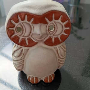 May include: A ceramic owl figurine in cream and brown. The owl has large, stylized eyes with sunburst details and a textured body. The figurine is a decorative item.