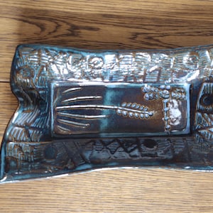 May include: A rectangular, handmade ceramic tray with a dark teal glaze. The tray features a central rectangular design with a wheat stalk motif and a decorative border. The tray is approximately 10 inches long and 6 inches wide.