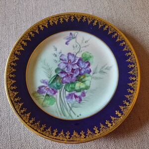 Może przedstawiać: Antyczny talerz porcelanowy z niebieską i złotą obwódką. Środek talerza zdobi ręcznie malowany wzór fioletowych fiołków z zielonymi liśćmi.