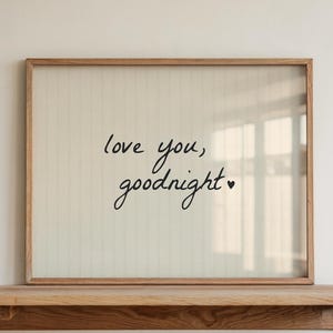 Op de afbeelding: Een ingelijste print met de woorden "love you, goodnight" in zwart schrift. De print zit in een licht houten frame en rust op een bijpassende houten plank. De achtergrond is een effen witte muur.