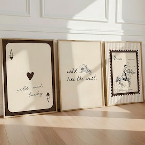 Könnte beinhalten: Drei gerahmte Kunstdrucke, die an einer weißen Wand lehnen. Die Drucke zeigen ein Spielkarten-Design, eine Cowboy-Illustration mit dem Text "wild like the west" und ein Vintage-Postwertzeichen-Design. Die Rahmen haben einen goldenen Rand.
